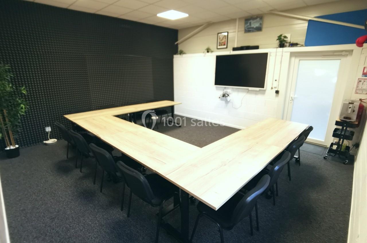 Salle de réunion avec tables en forme de U, chaises noires, écran mural et éclairage au plafond.