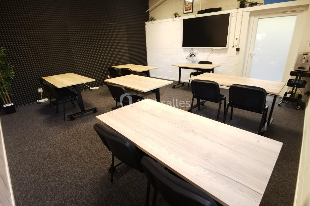 Salle de réunion équipée de tables en bois clair, chaises noires, écran mural et murs insonorisés.