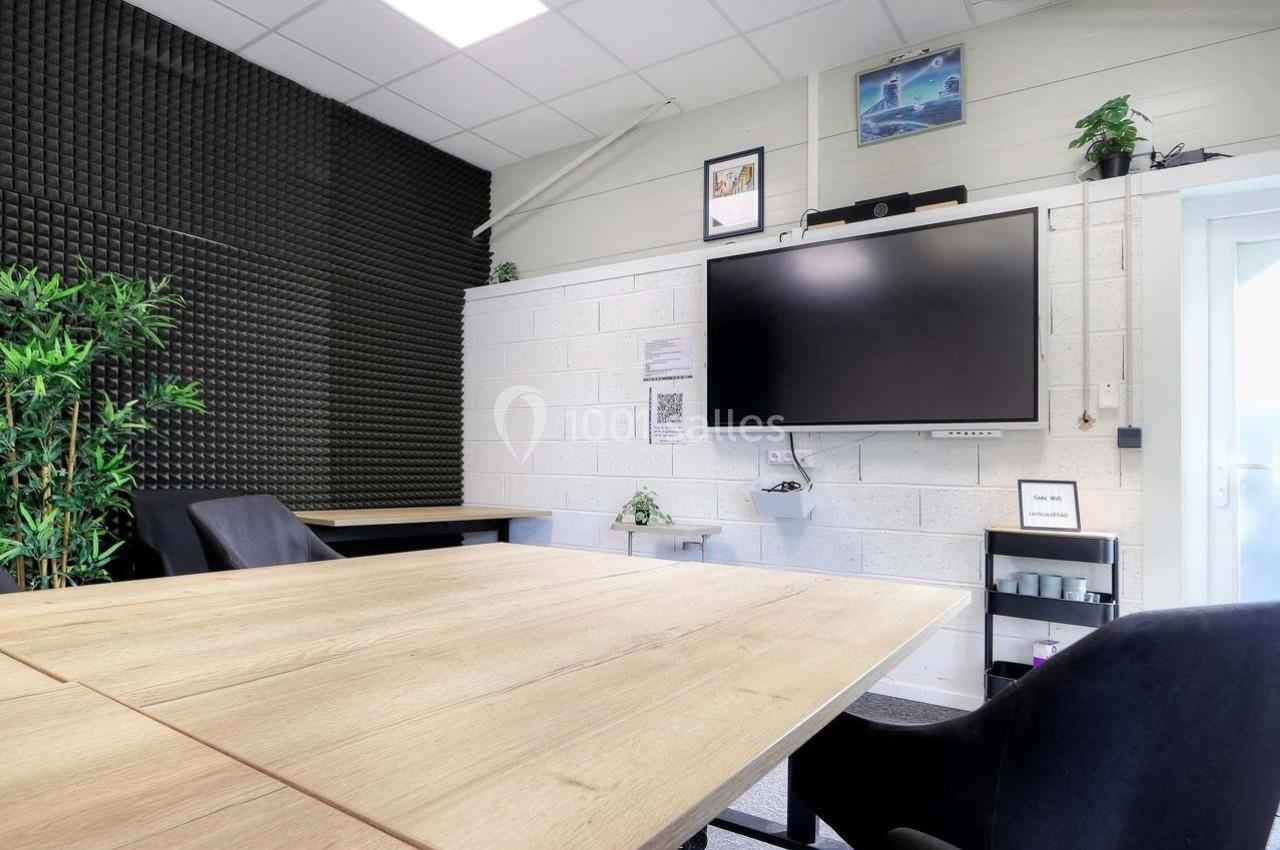 Salle de réunion moderne avec table en bois, chaises noires, écran mural et mur partiellement insonorisé.