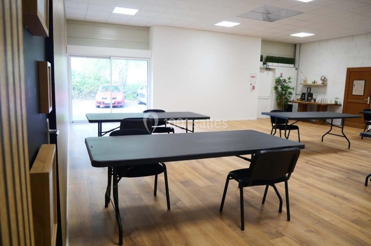Salle lumineuse avec tables noires espacées, chaises, sol en bois, et une baie vitrée donnant sur l'extérieur.