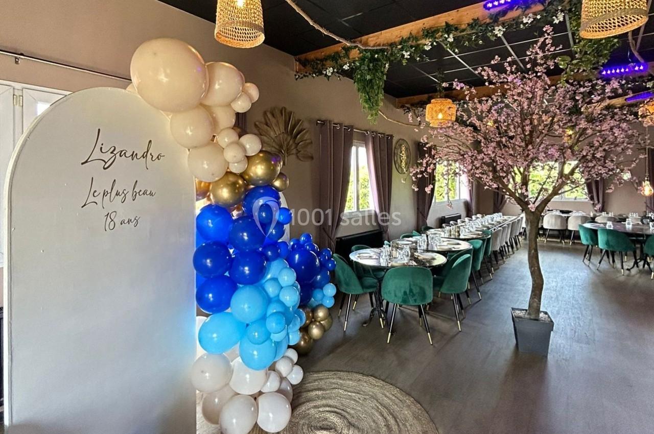 Salle décorée pour un événement avec ballons bleus et blancs, table dressée et arbre artificiel fleuri.