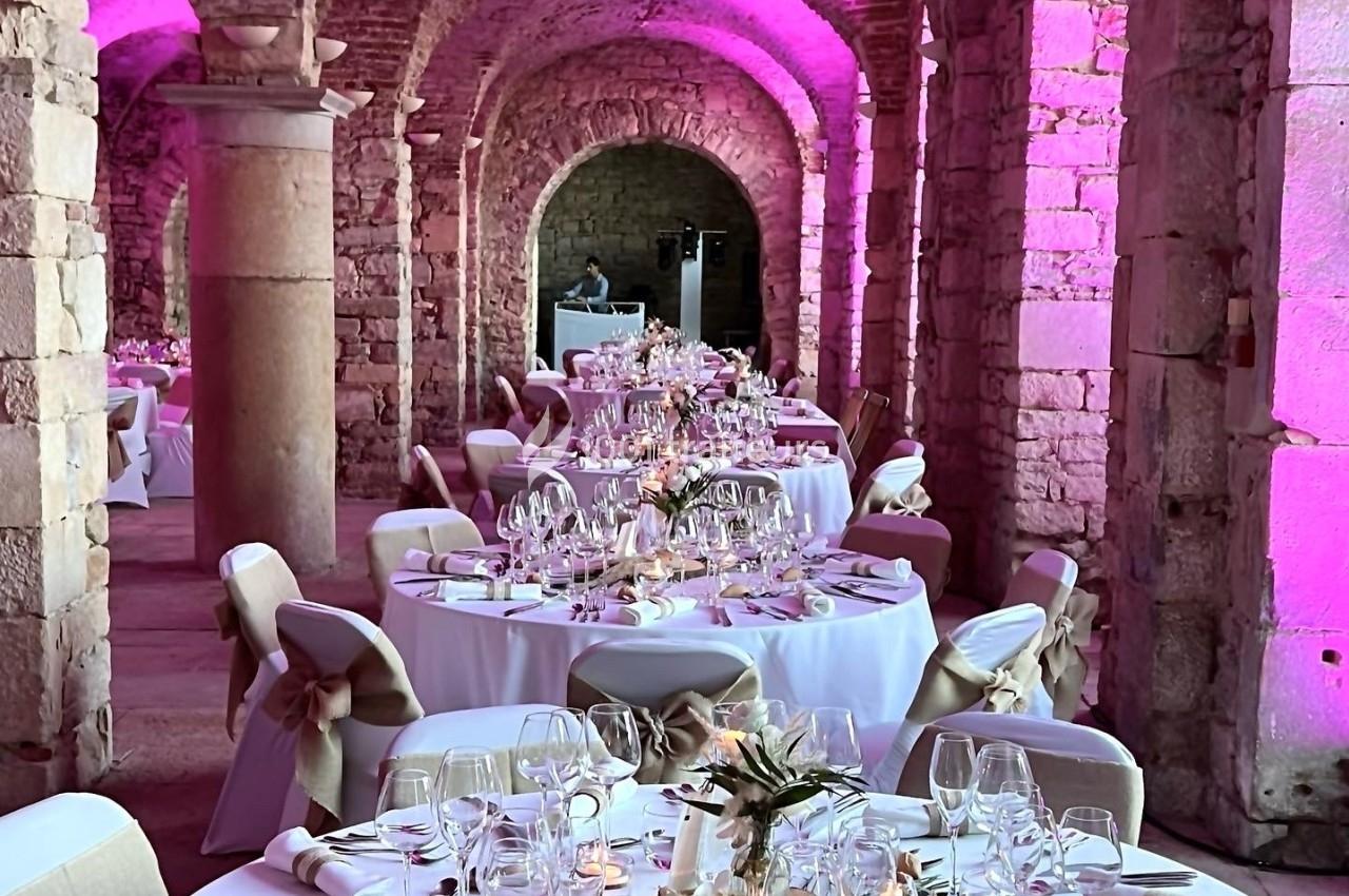 Salle voûtée en pierre décorée pour un mariage, avec tables rondes dressées et éclairage rose.