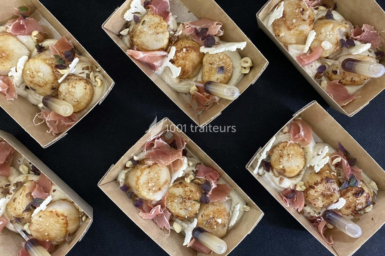 Coquilles Saint-Jacques poêlées avec jambon cru, noisettes concassées et pipettes de sauce, présentées en barquettes.