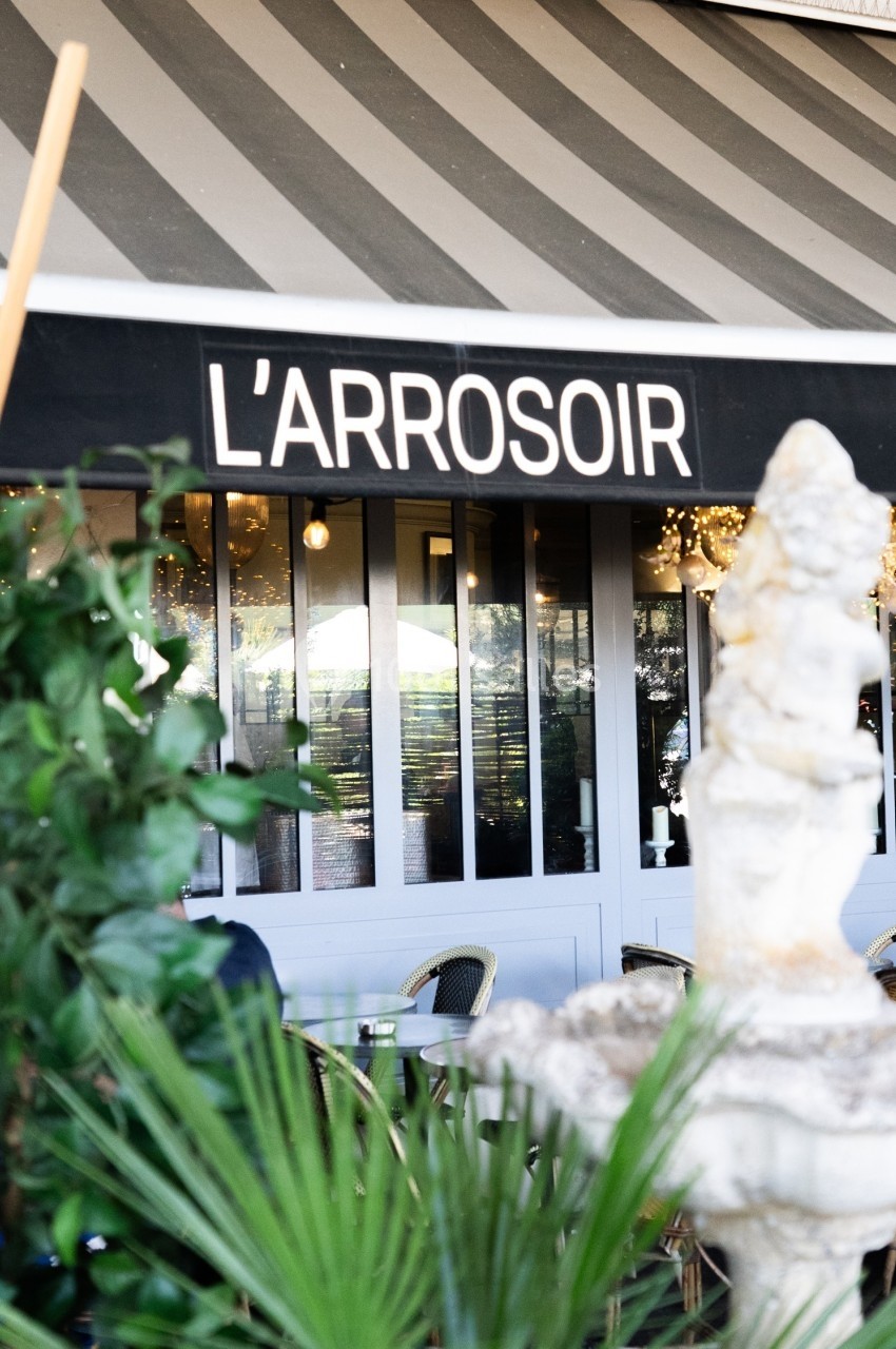 Façade d'un restaurant nommé ’L'Arrosoir’ avec une terrasse partiellement visible et des plantes au premier plan.