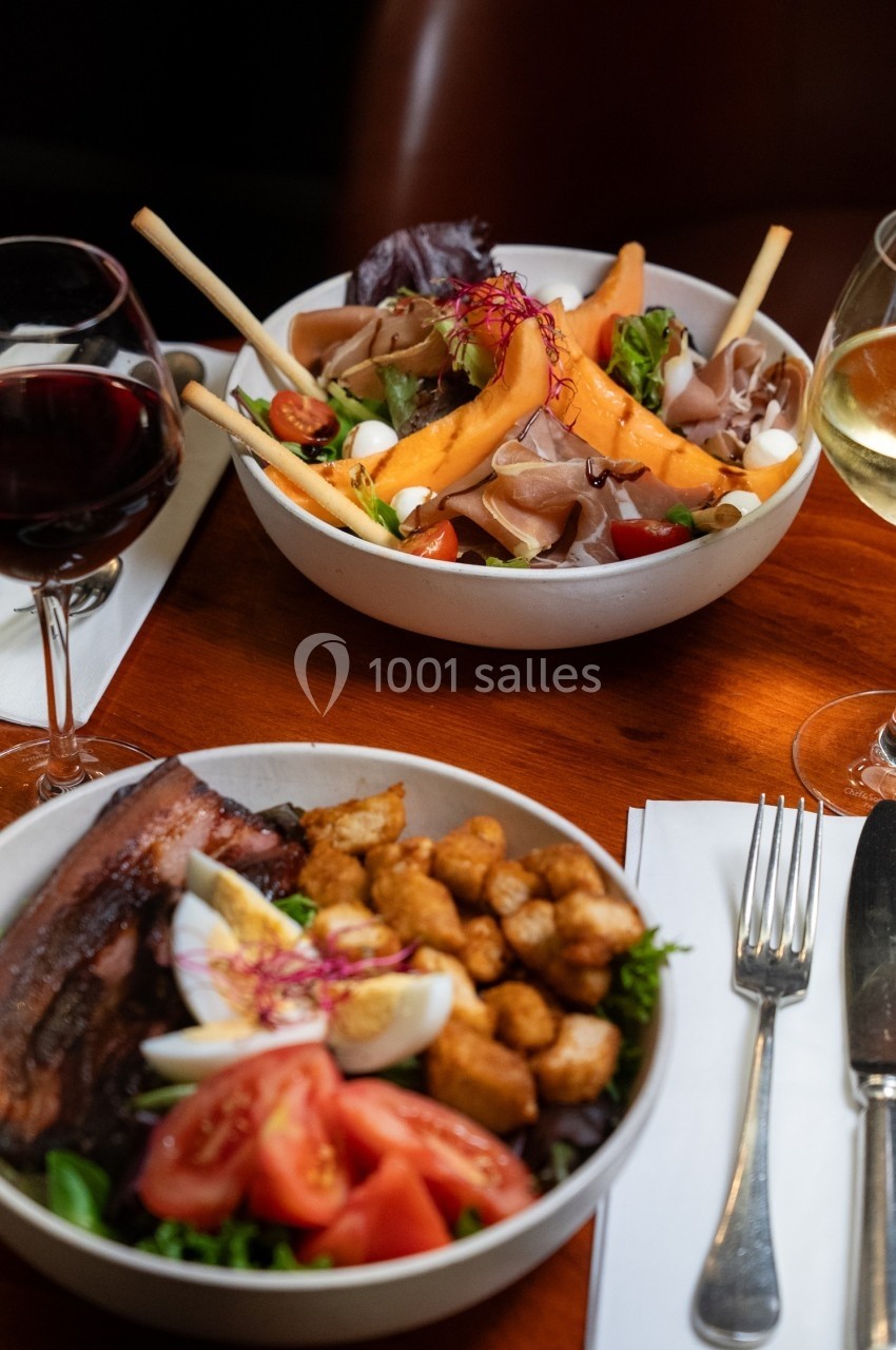 Deux assiettes garnies de salades variées avec légumes, viande et fromage, accompagnées de verres de vin rouge et blanc.