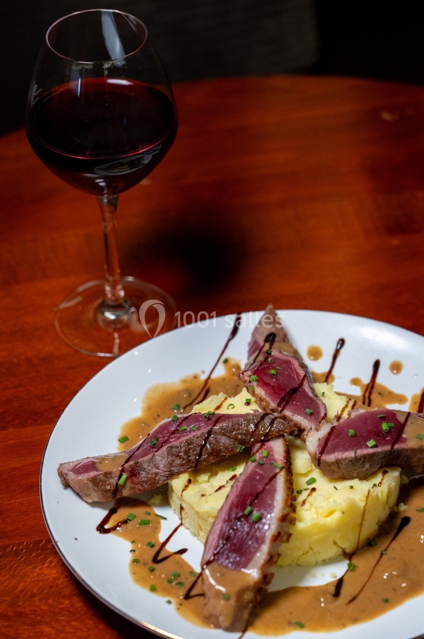 Assiette de magret de canard rosé sur purée, nappée de sauce, avec un verre de vin rouge en arrière-plan.