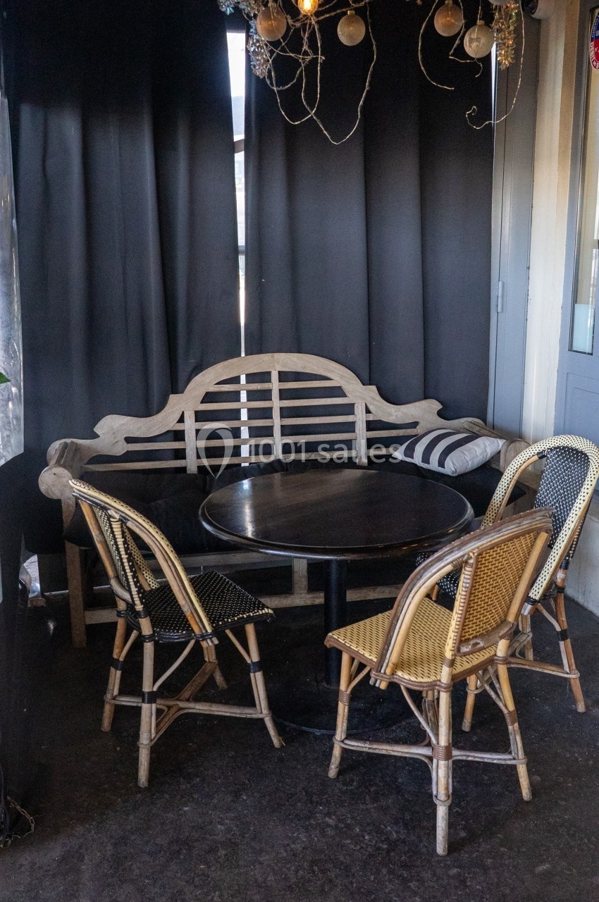 Table ronde en bois entourée de chaises en rotin et d'un banc, dans un espace intérieur avec rideaux sombres.