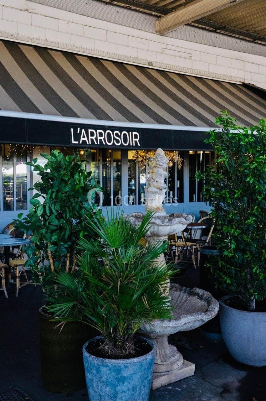 Devanture d'un café-restaurant avec auvent rayé, plantes en pots et une fontaine décorative au premier plan.