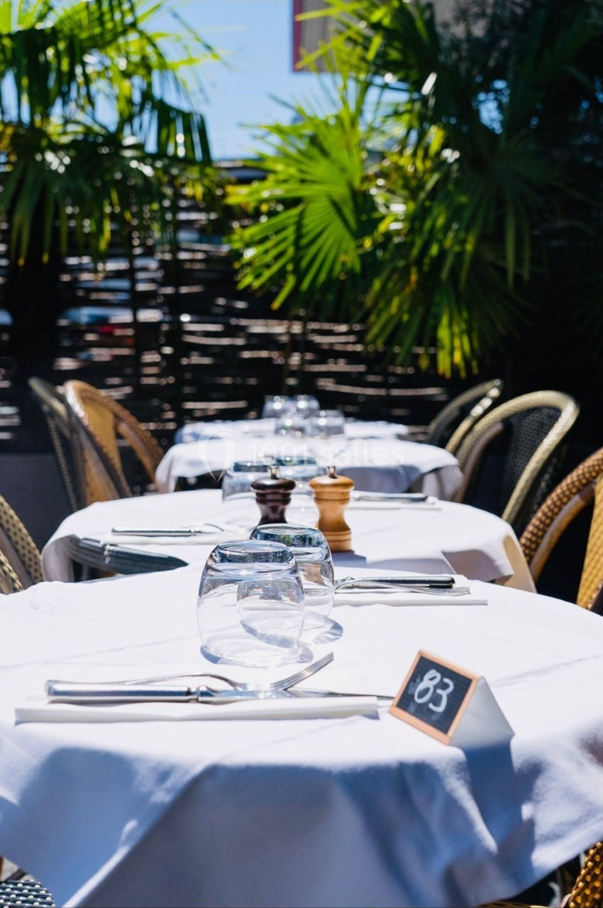 Tables de restaurant en extérieur avec nappes blanches, couverts, verres et numéro de table, entourées de plantes tropicales.