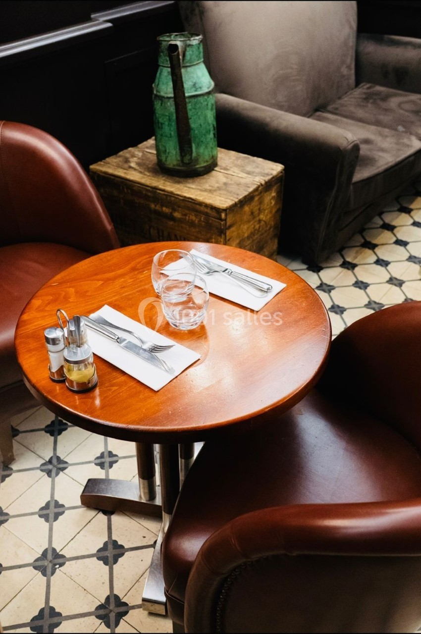 Table ronde en bois avec deux couverts, un verre d'eau et des condiments, entourée de fauteuils en cuir marron.