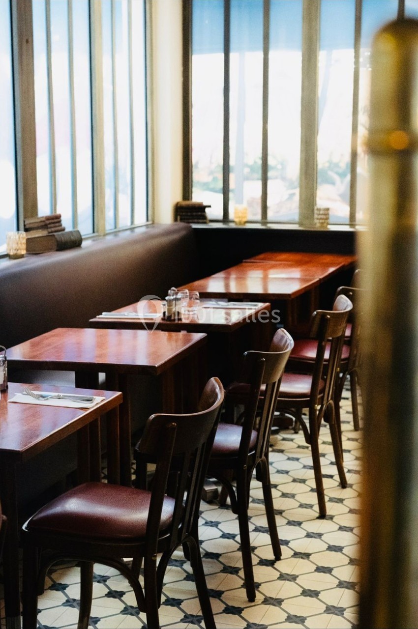 Salle de restaurant avec tables en bois, chaises assorties et lumière naturelle entrant par de grandes fenêtres.