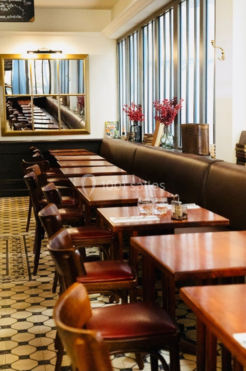 Salle de restaurant lumineuse avec tables en bois, chaises en cuir, carrelage au sol et vase de fleurs rouges sur une table.