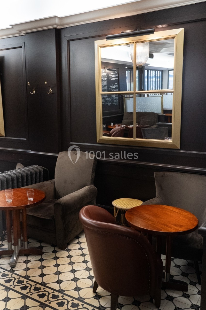 Deux fauteuils et deux tables en bois dans un salon au décor sombre avec un miroir encadré doré au mur.