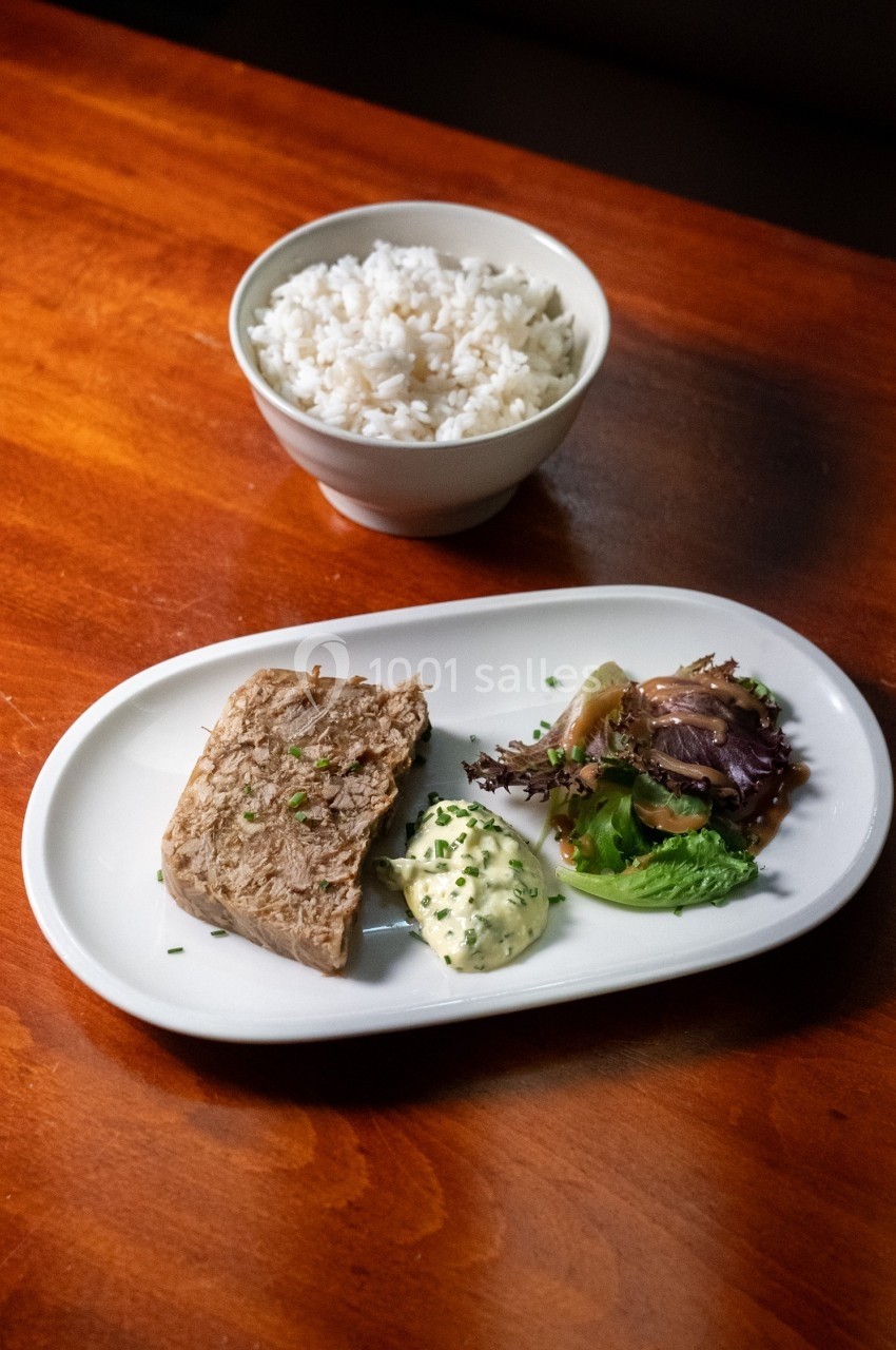 Assiette contenant une portion de viande, légumes, sauce et herbes, accompagnée d'un bol de riz blanc en arrière-plan.