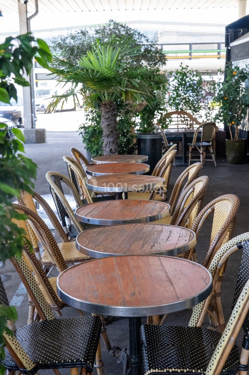 Terrasse extérieure avec des tables rondes en bois et des chaises en osier, entourées de plantes vertes.