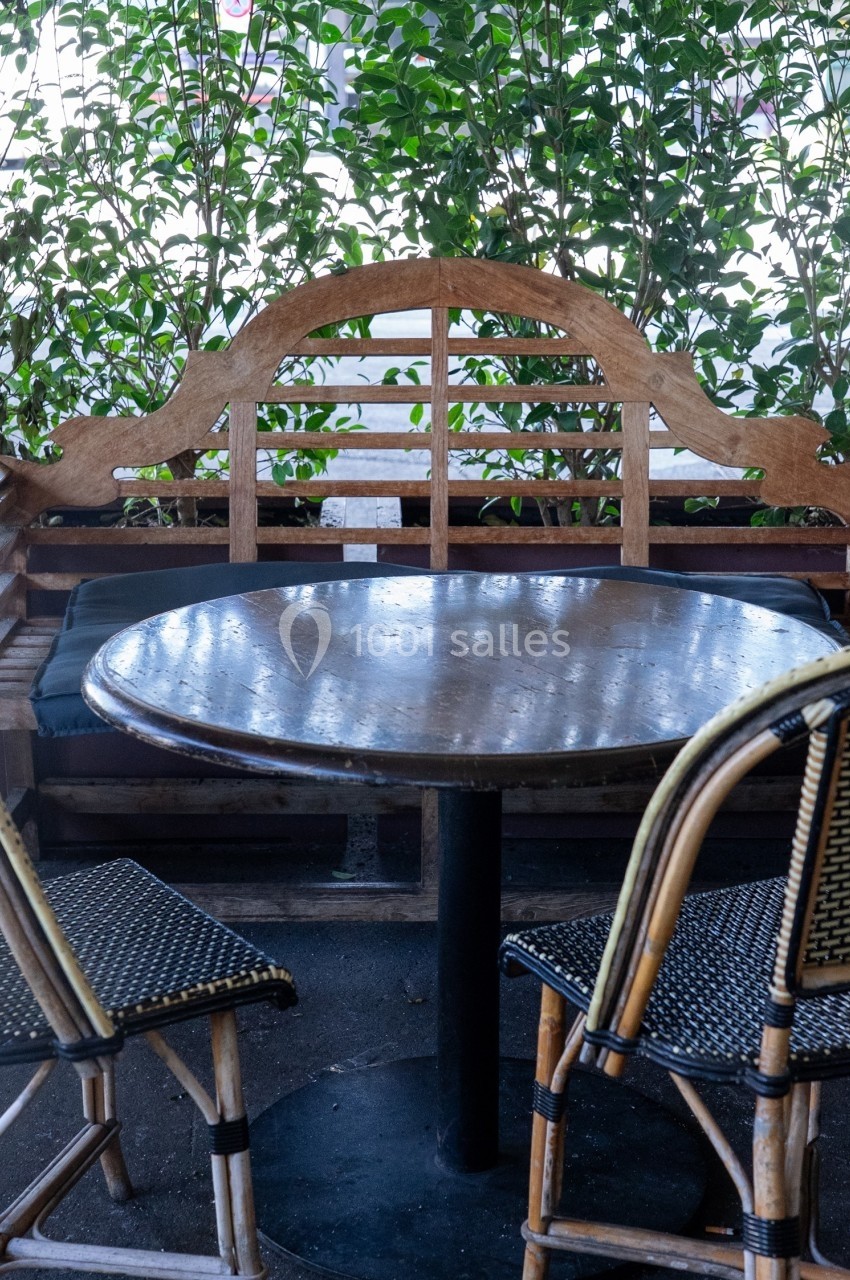 Table ronde en bois entourée de deux chaises en osier et d'un banc, située sur une terrasse avec des plantes en arrière-plan.
