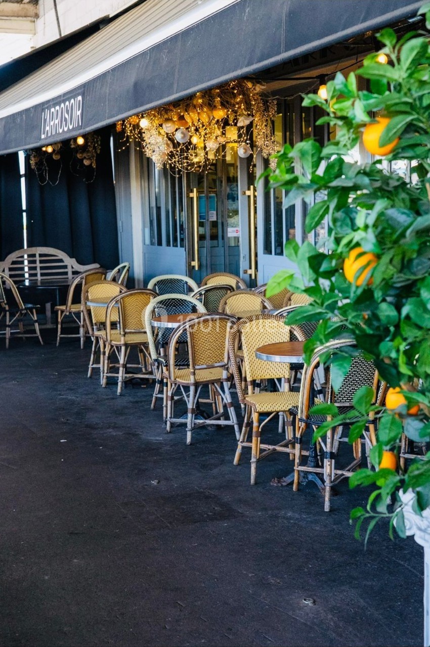 Terrasse d'un café avec des chaises en osier, une enseigne ’L'Approsoir’ et un oranger en premier plan.