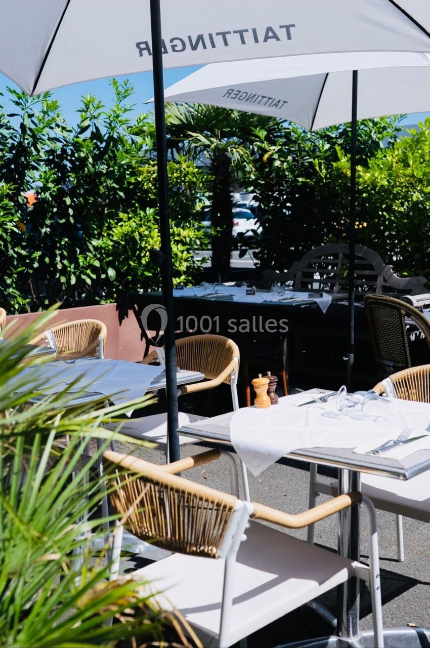 Terrasse extérieure avec tables dressées, chaises en rotin et parasols blancs, entourée de verdure.