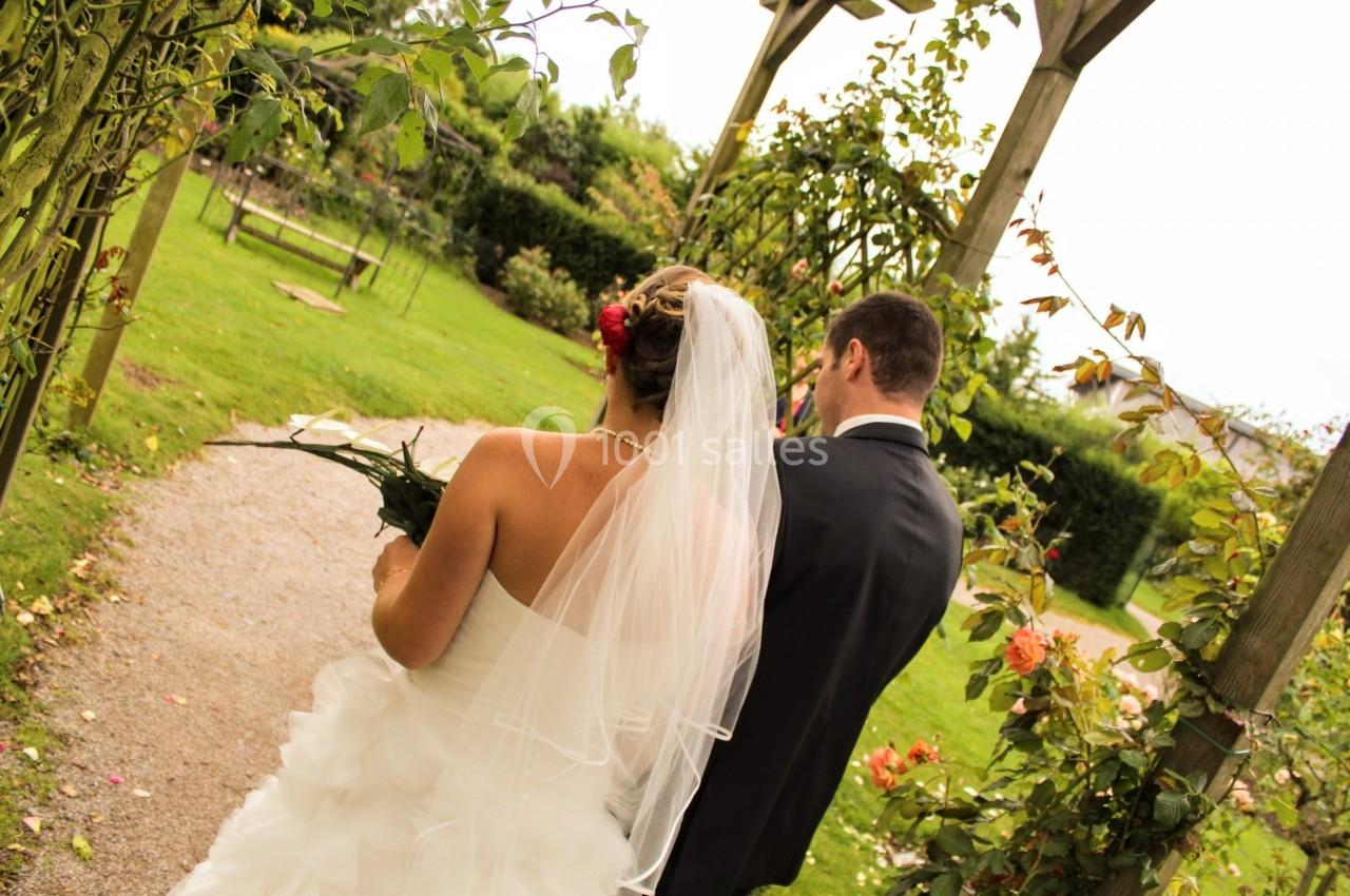 Un couple de mariés marchant dans un jardin sous une pergola, entouré de verdure et de fleurs.