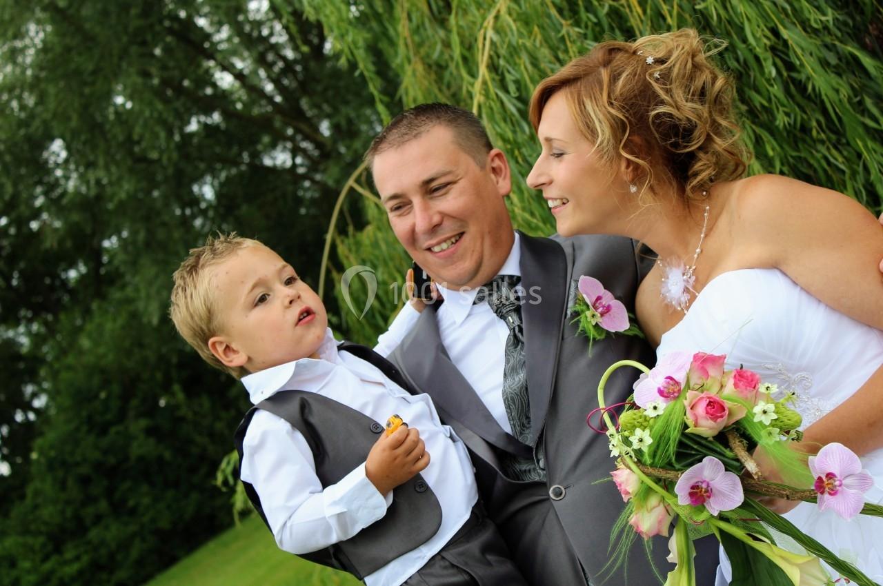 Un couple en tenue de mariage sourit en portant un jeune garçon, avec un fond de verdure et de saules pleureurs.