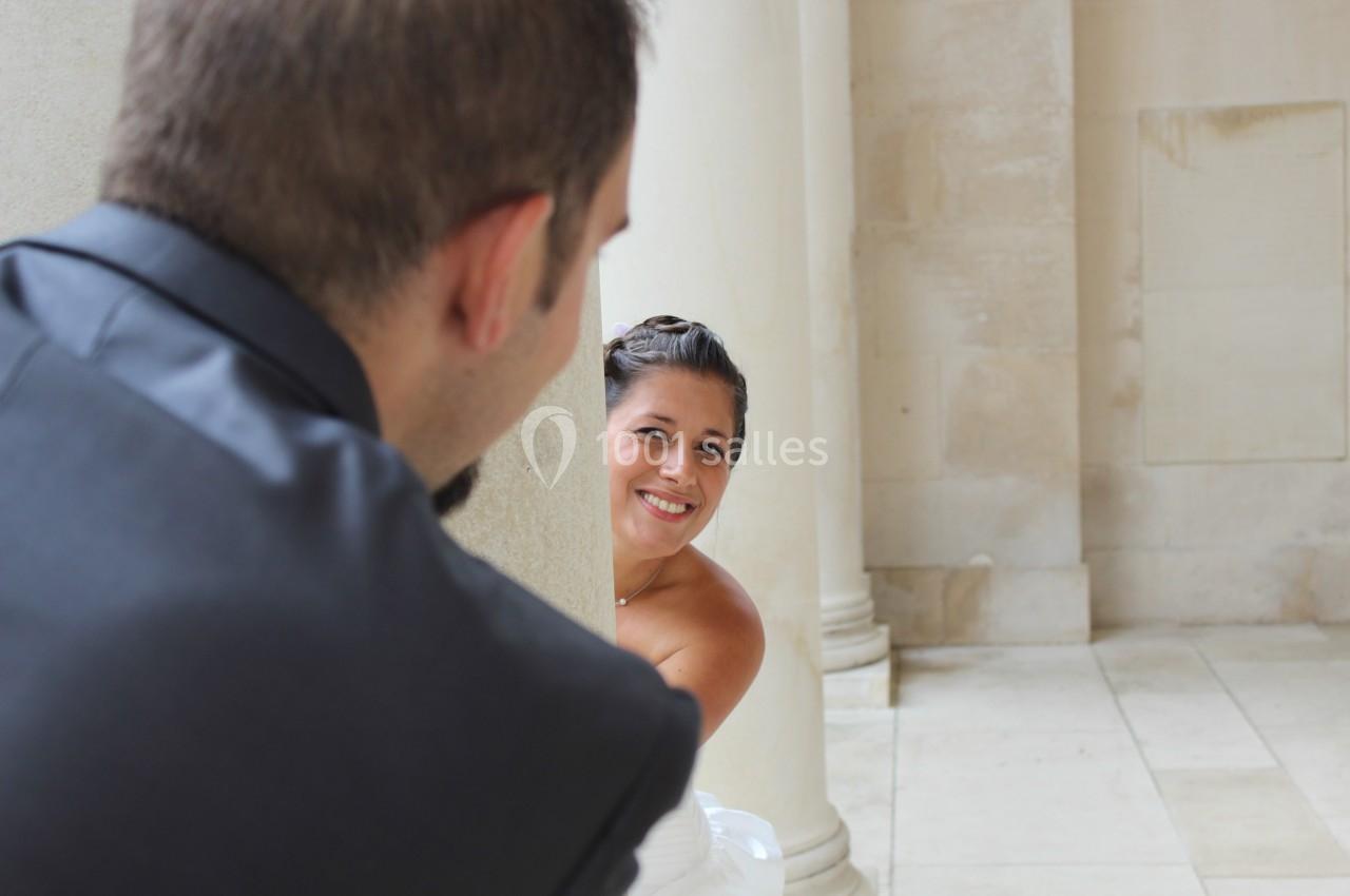Une femme souriante regarde derrière une colonne, face à un homme de dos dans un cadre architectural clair.