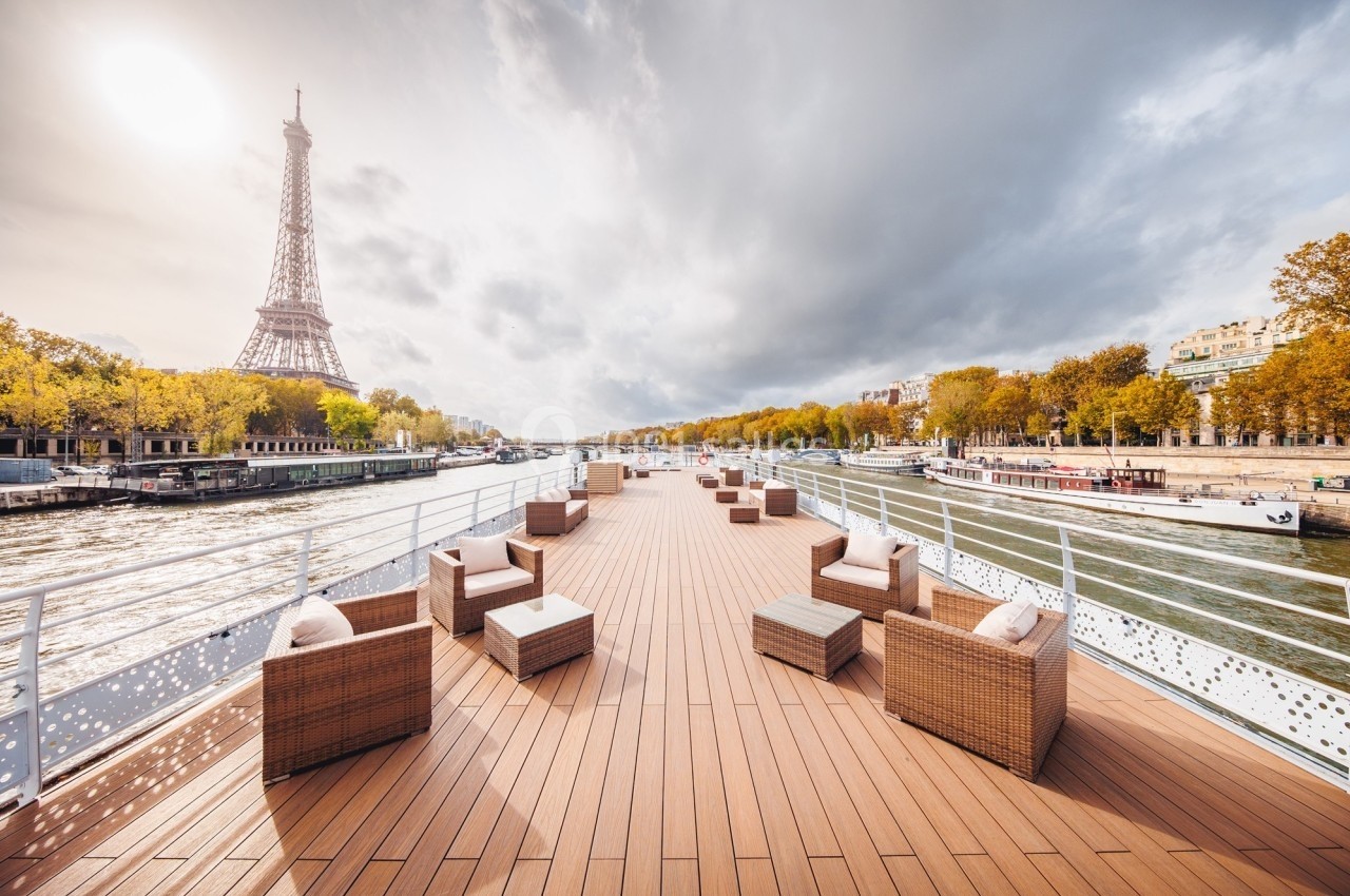 Pont d'un bateau avec mobilier en rotin, vue sur la Seine et la tour Eiffel sous un ciel partiellement nuageux.