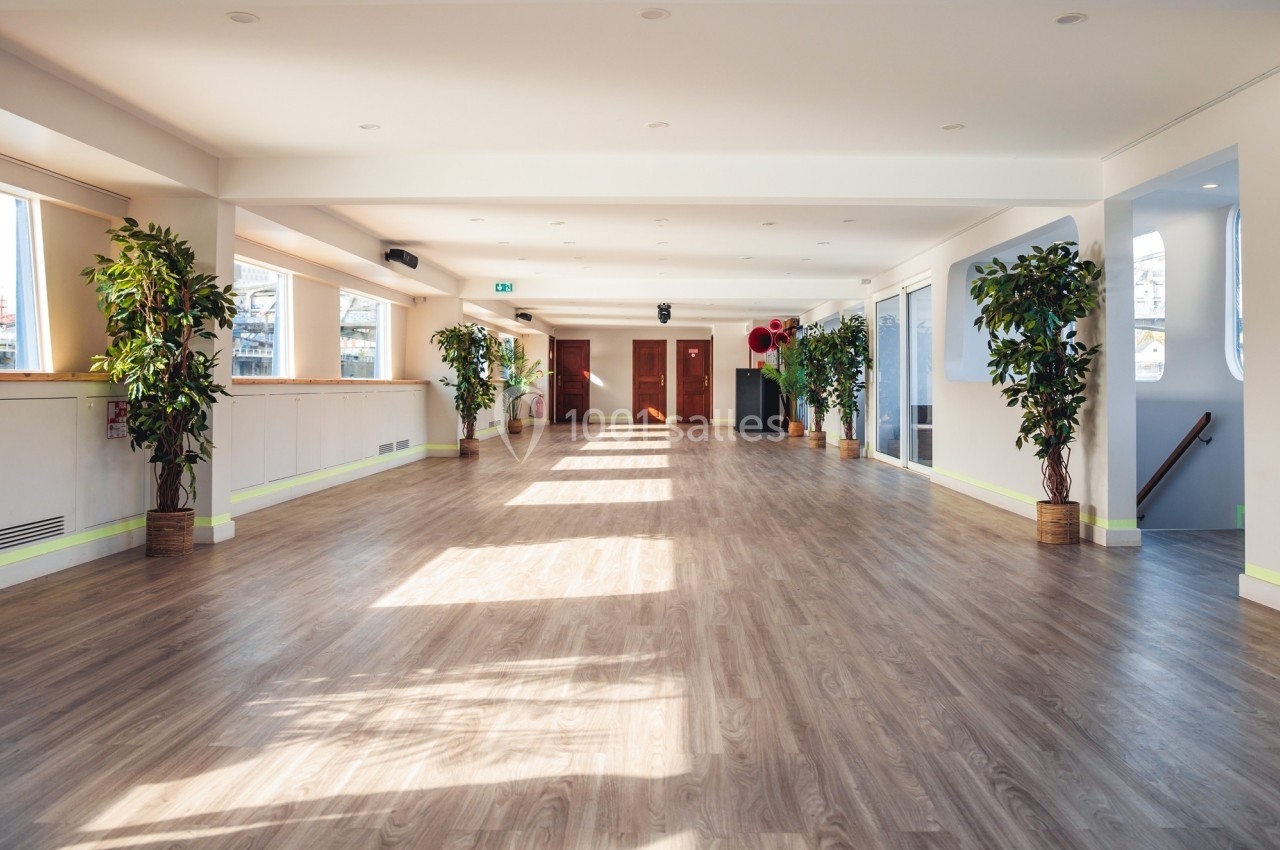 Salle lumineuse avec parquet, grandes fenêtres laissant entrer la lumière naturelle, et plantes disposées le long des murs.
