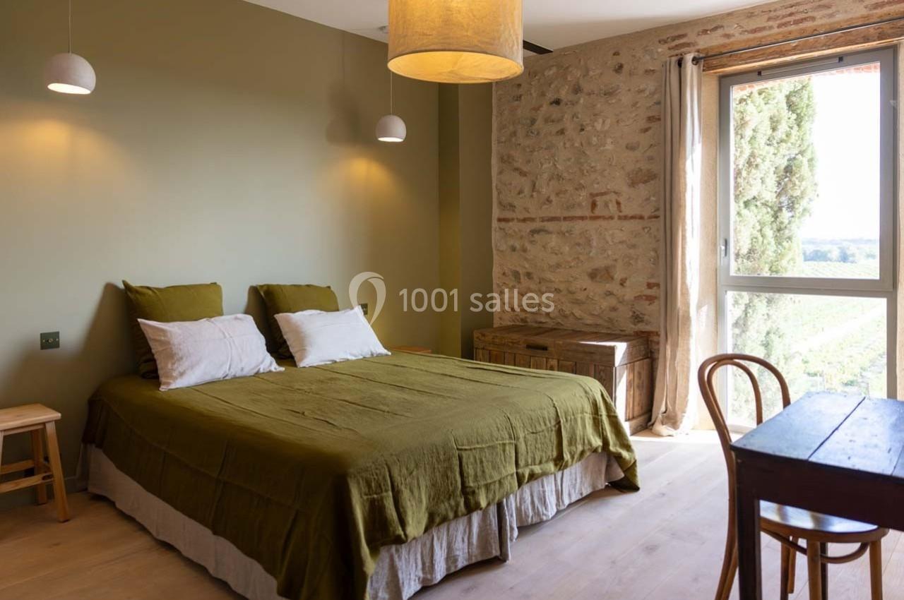 Chambre lumineuse avec lit double, murs en pierre et vue sur la campagne depuis une grande fenêtre.