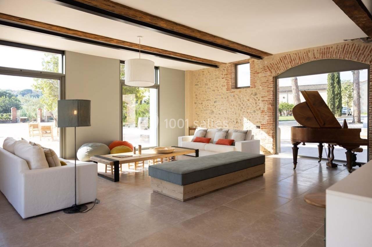 Salon lumineux avec murs en pierre, mobilier moderne, piano à queue et grandes baies vitrées donnant sur l'extérieur.