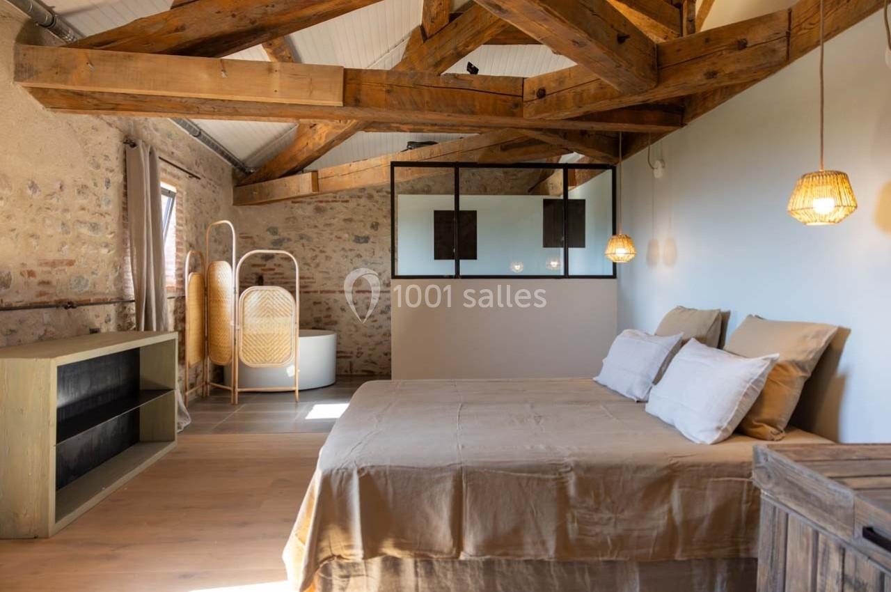 Chambre lumineuse avec lit double, poutres apparentes, murs en pierre, verrière et espace salle de bain en arrière-plan.