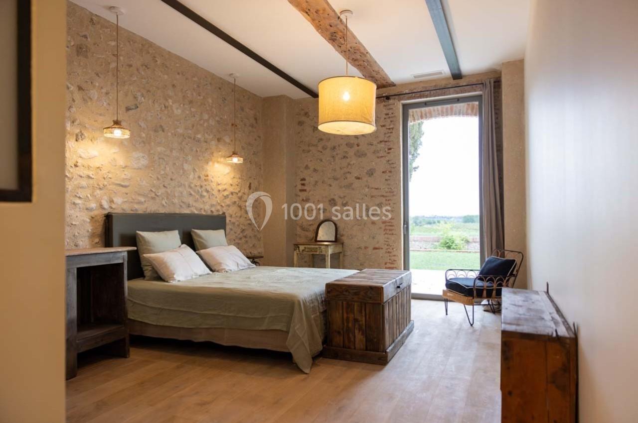Chambre lumineuse avec murs en pierre, lit double, mobilier en bois, et vue sur un paysage extérieur par une porte vitrée.