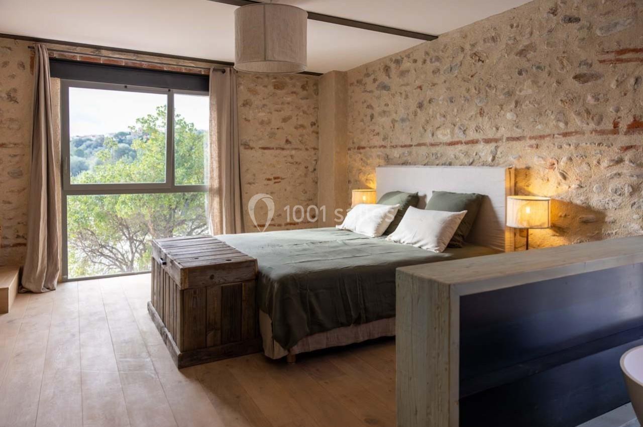 Chambre lumineuse avec murs en pierre, lit double, coffre en bois, grandes fenêtres et vue sur la nature.