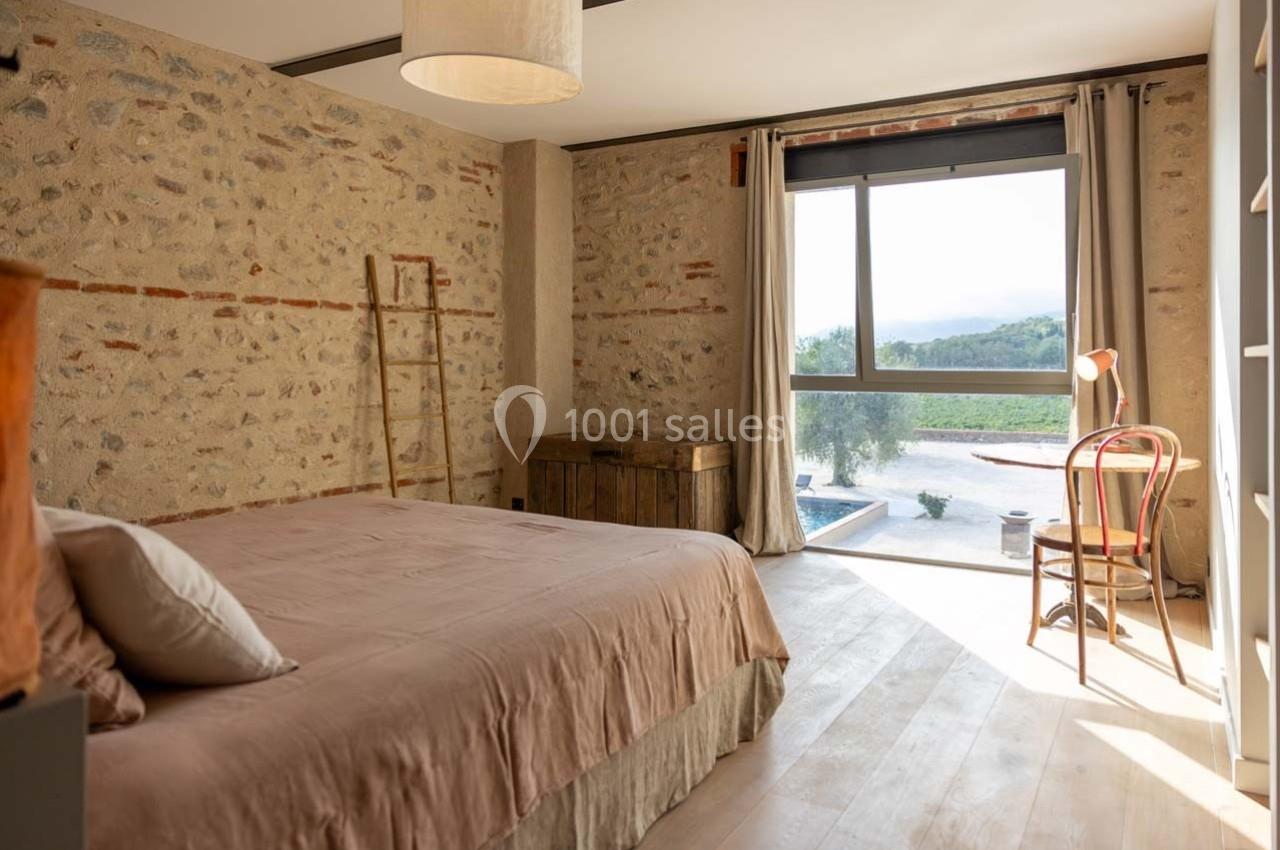 Chambre lumineuse avec murs en pierre, lit double, chaise en bois, fenêtre donnant sur un paysage naturel.