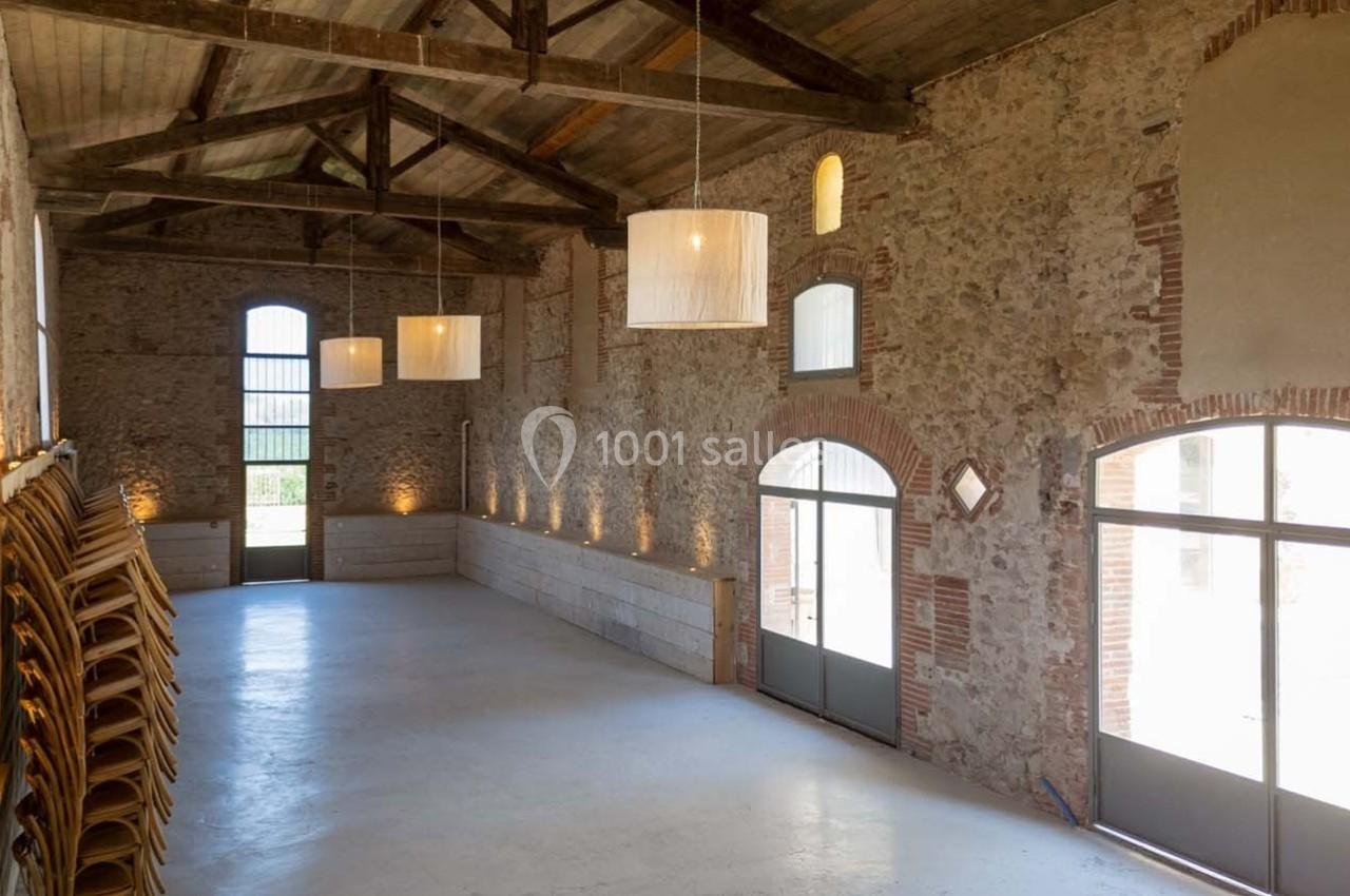 Salle en pierre avec charpente en bois, grandes fenêtres cintrées, suspensions lumineuses et chaises empilées sur le côté.