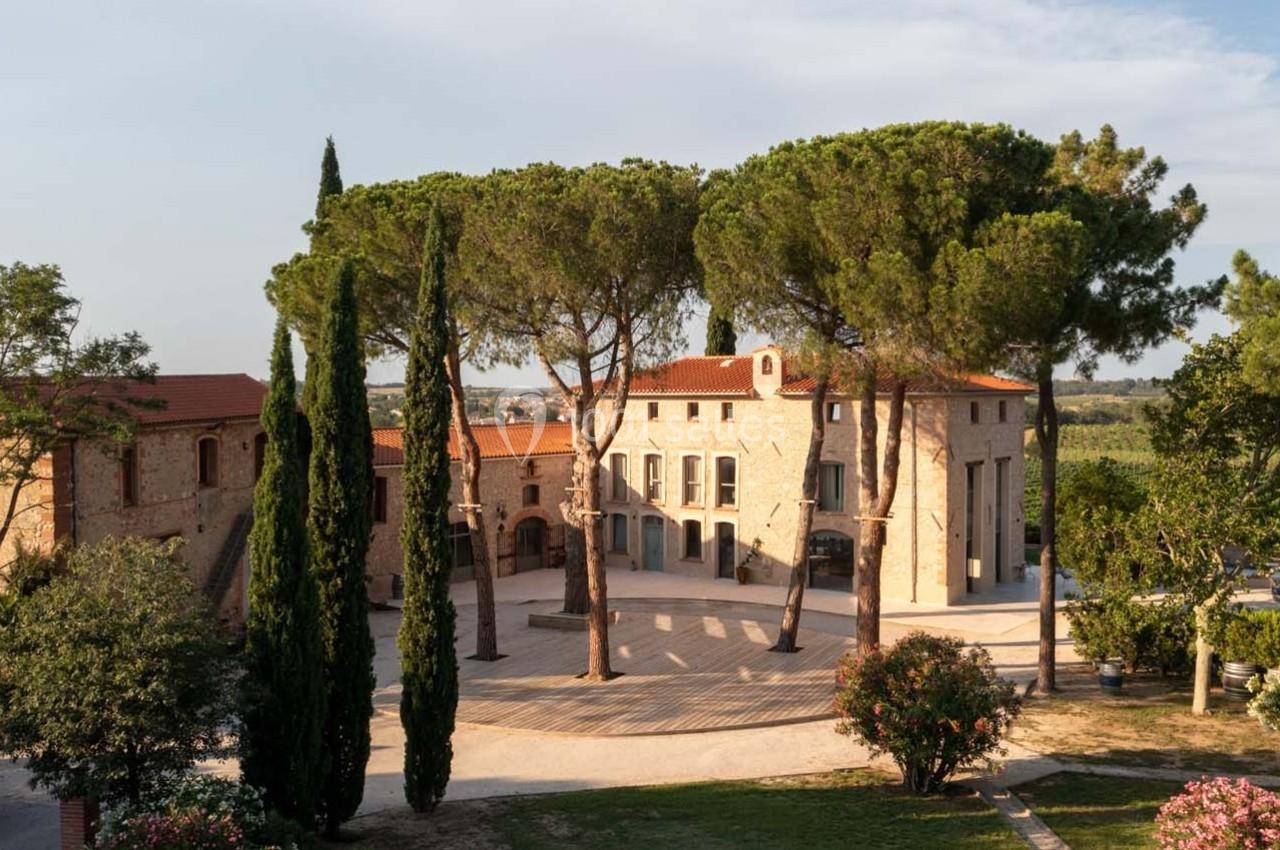Vue d'un domaine avec bâtiments en pierre, arbres méditerranéens et cour pavée sous un ciel dégagé.