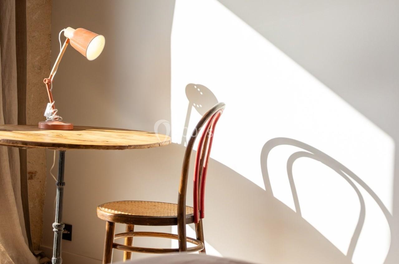 Lampe de bureau allumée sur une table en bois, chaise en osier et ombre projetée sur un mur clair.