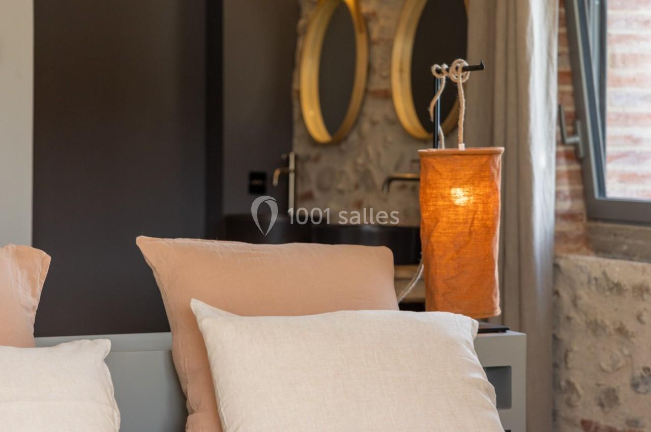 Lit avec oreillers beiges, lampe allumée sur une table de chevet, mur en pierre et miroirs en arrière-plan.