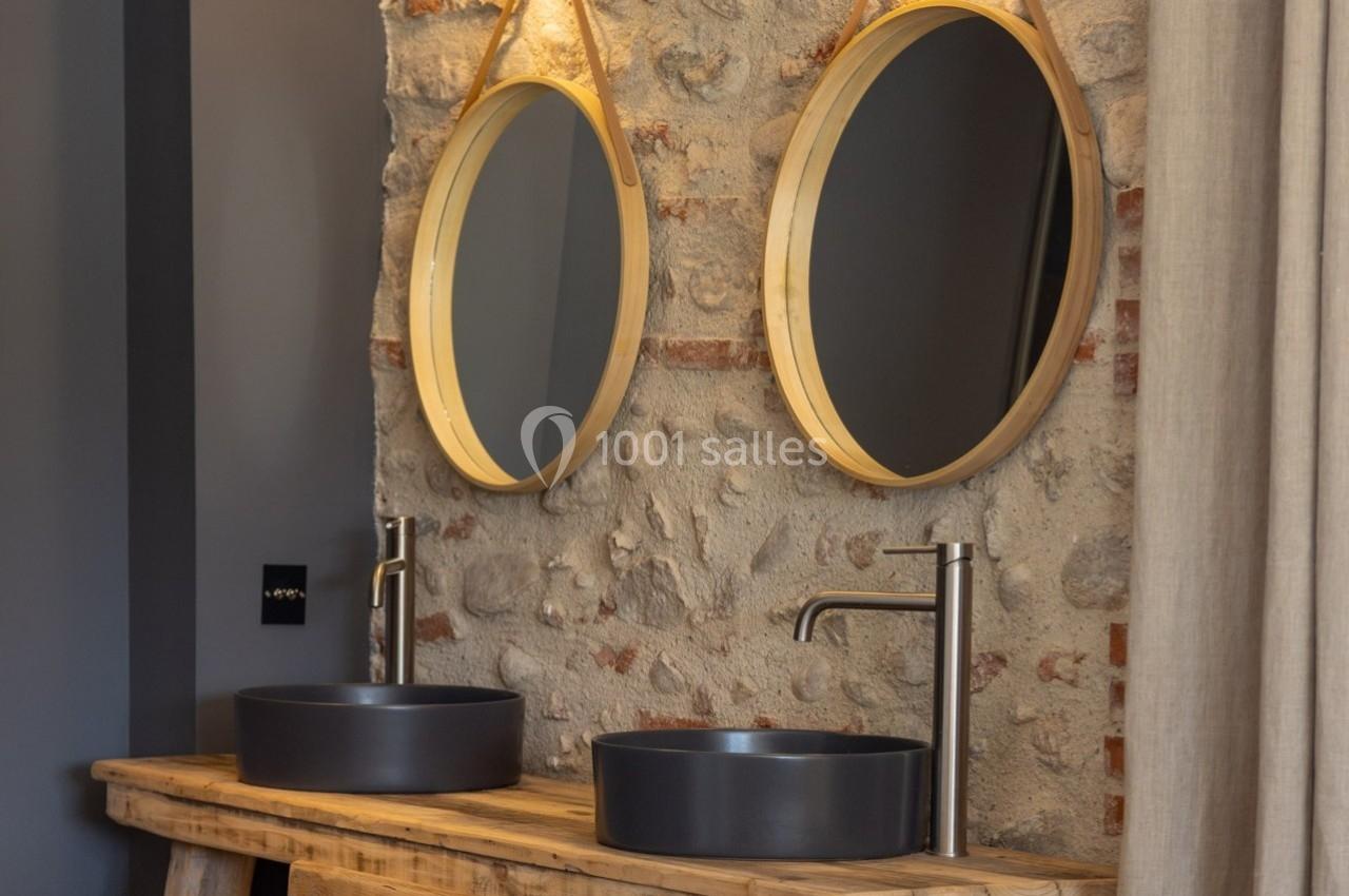 Deux lavabos noirs avec robinets modernes sur un meuble en bois, devant un mur en pierre avec deux miroirs ronds.