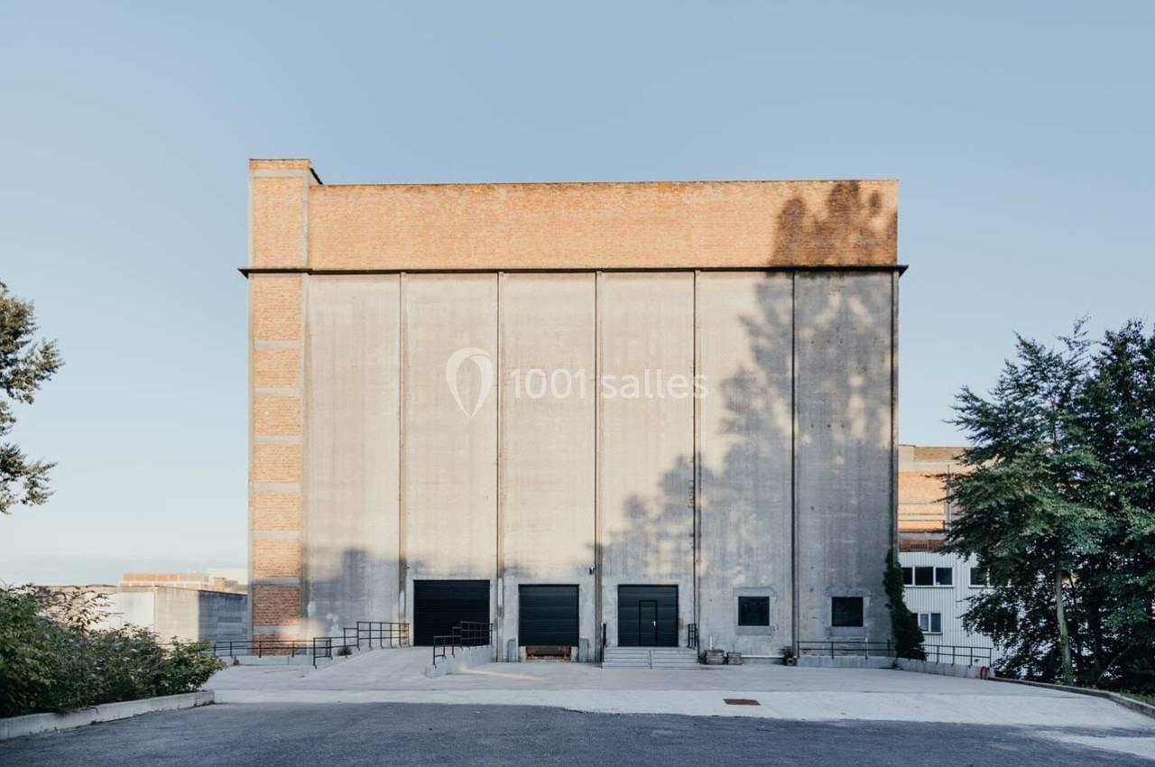 Bâtiment industriel en briques et béton avec trois grandes portes noires, entouré d'arbres et d'une cour pavée.