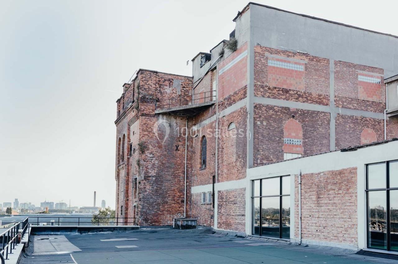 Bâtiment en briques rouges avec des fenêtres modernes, situé sur un toit-terrasse avec vue sur une ville à l'arrière-plan.