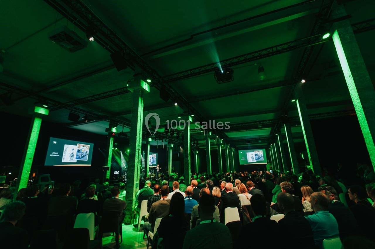 Salle de conférence avec un public assis, éclairage vert et écrans projetant des présentations.