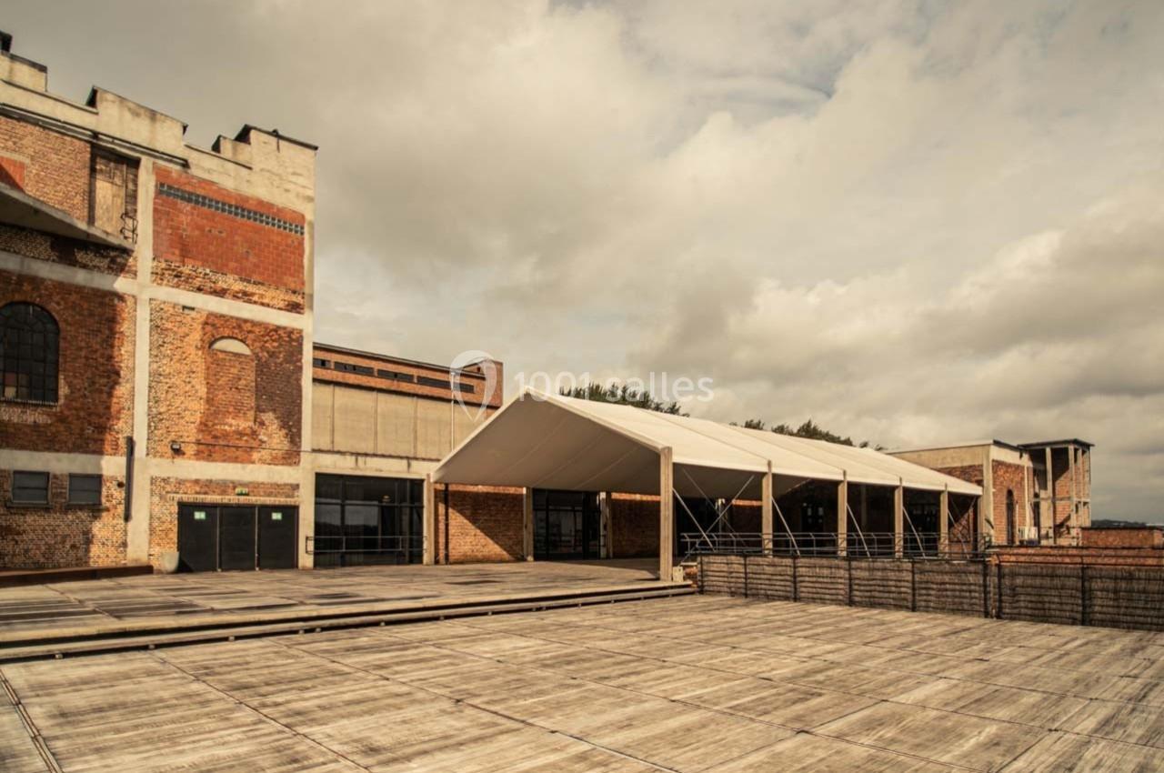 Bâtiment en briques rouges avec une structure moderne en toile blanche sur une grande esplanade en bois.