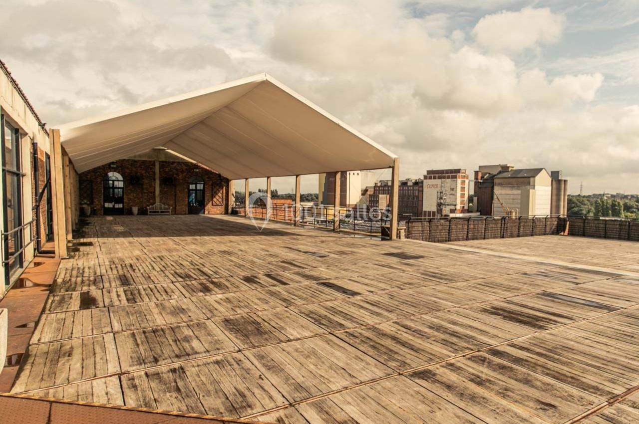 Terrasse en bois spacieuse avec une structure couverte, entourée de bâtiments urbains sous un ciel partiellement nuageux.