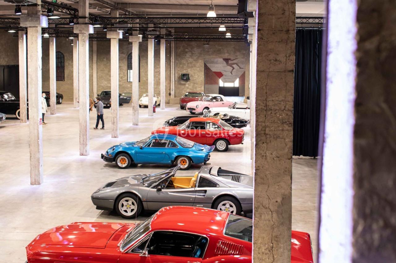 Collection de voitures classiques et sportives exposées dans un grand espace industriel avec des colonnes en béton.