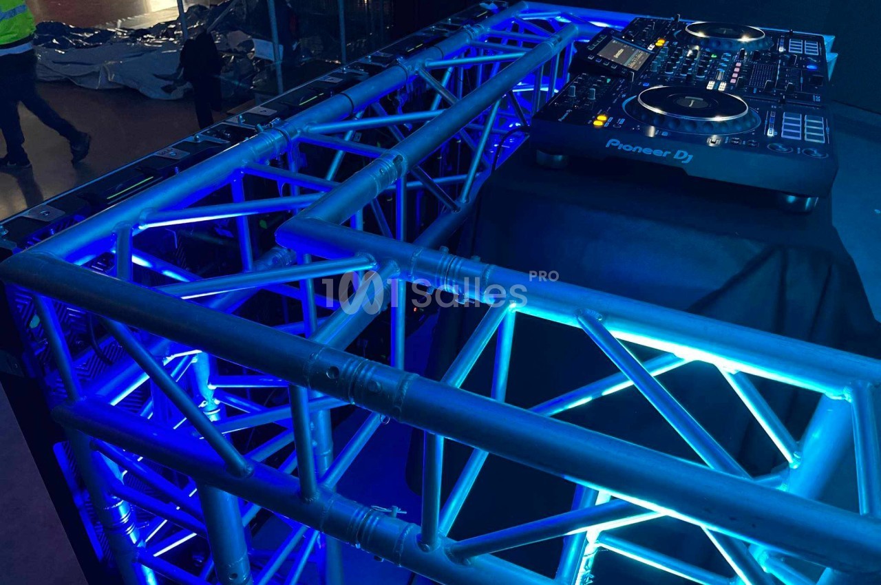 Structure métallique éclairée en bleu avec une table de mixage DJ posée dessus, dans un environnement sombre.