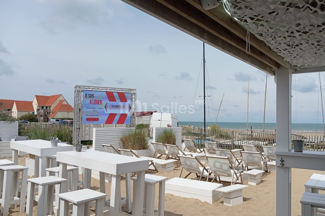 Terrasse de plage avec tables et chaises blanches, écran publicitaire, vue sur la mer et maisons en arrière-plan.