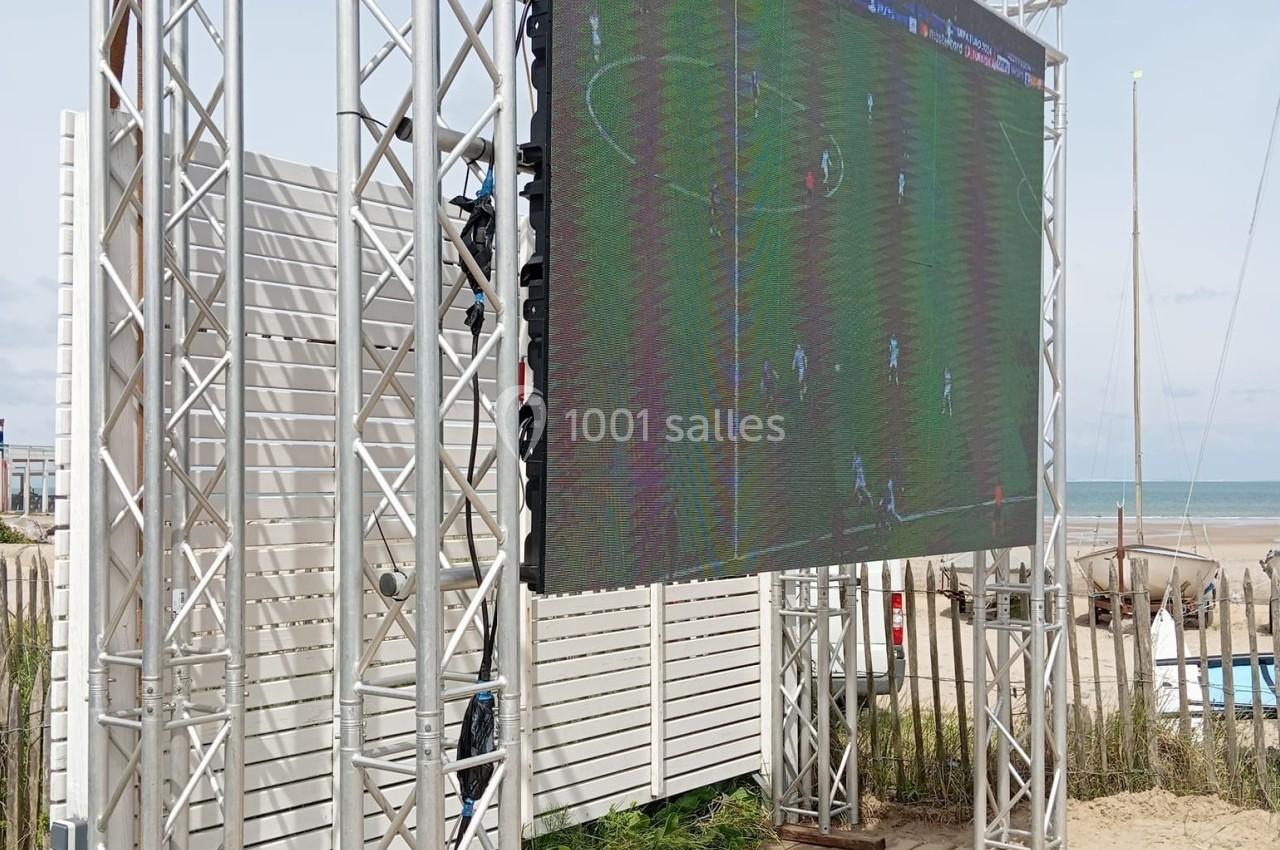 Écran géant installé sur une plage, diffusant un match de football, entouré de structures métalliques.