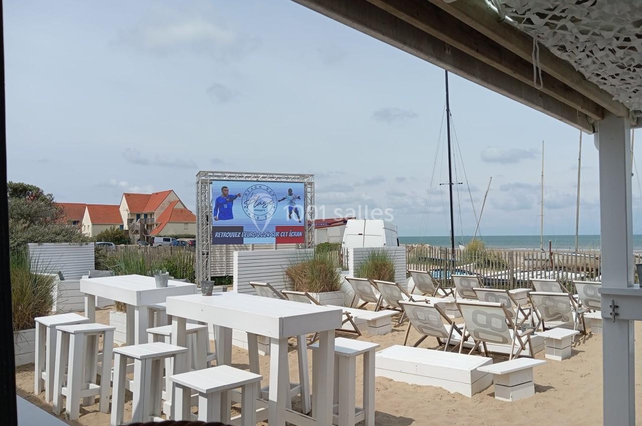 Terrasse en bord de mer avec tables et chaises blanches, vue sur la plage, la mer et un écran affichant une publicité.