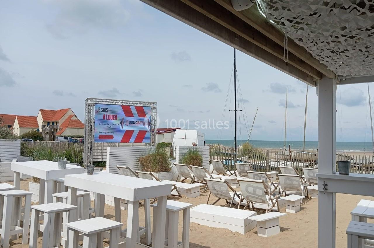 Terrasse de plage avec chaises longues, tables blanches et vue sur la mer, sous un ciel légèrement nuageux.