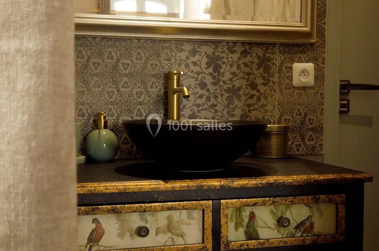 Lavabo noir sur meuble décoré d'oiseaux, robinet doré, carrelage à motifs et miroir encadré en arrière-plan.